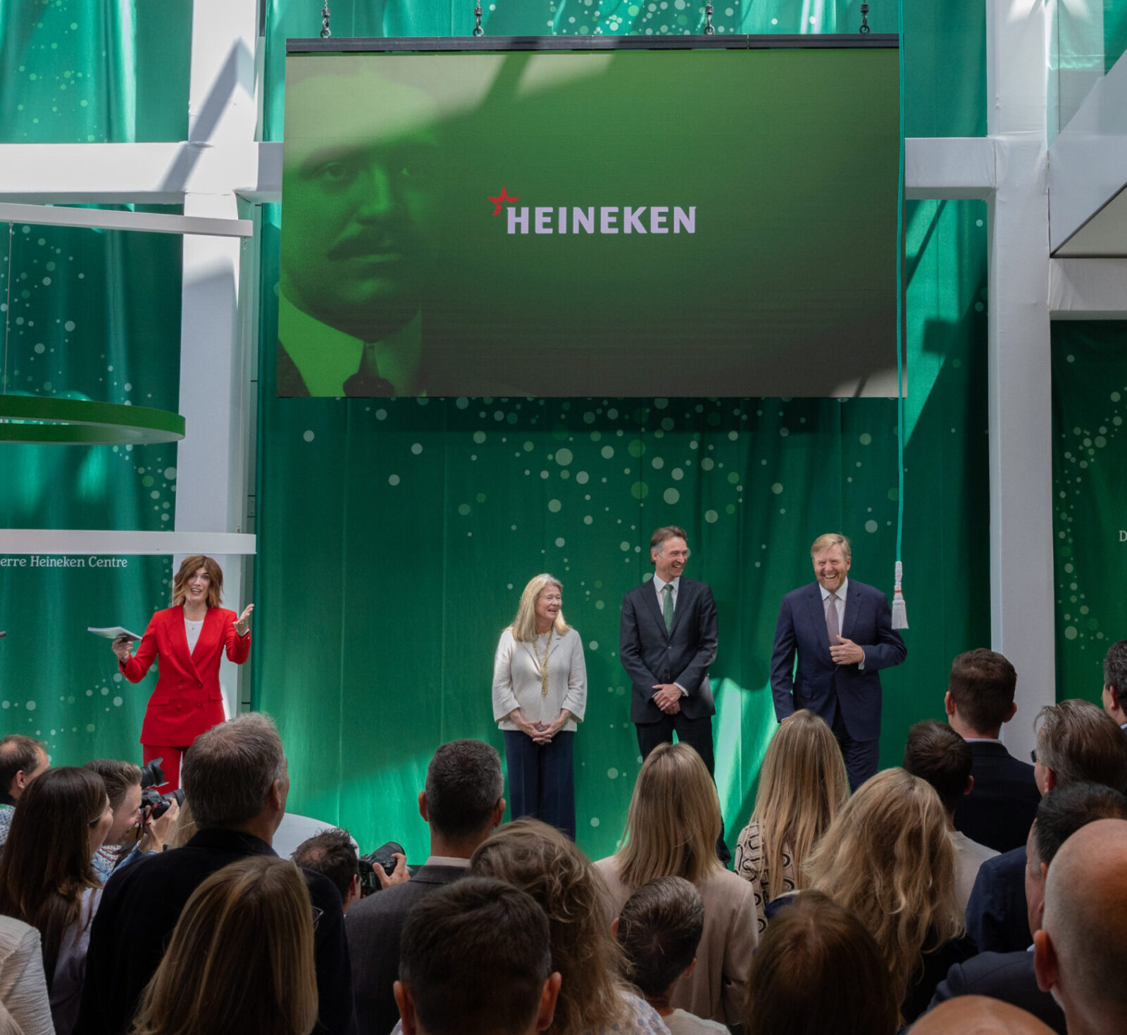 Koning Willem Alexander op het podium met Dolf van den Brink. Mevrouw De Carvalho-Heineken en Demelza van der Maas, bij de opening van het nieuwe R&D Cenytre in Zoeterwoude, met boven hun hoofd een groot scherm met daarop Henry Pierre Heineken afgebeeld.